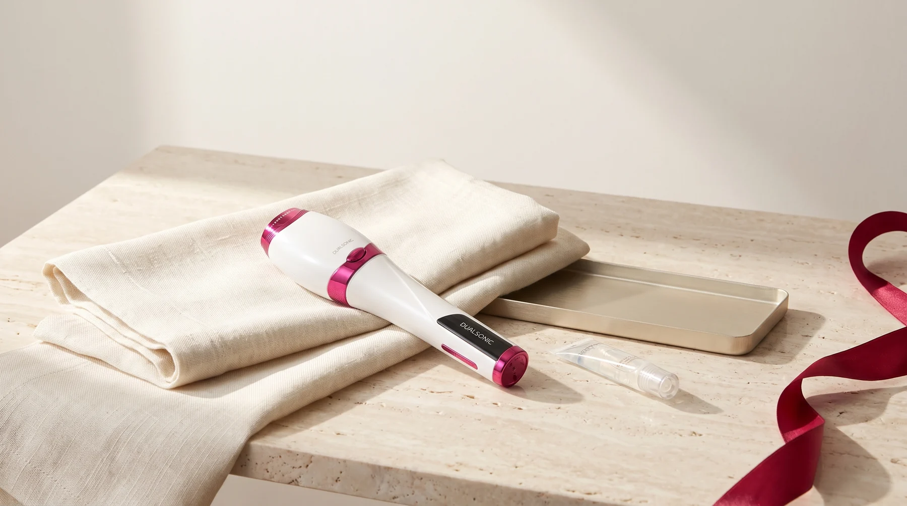Dualsonic Optimum posé sur du lin plié à côté d'un ruban rubis et d'un tube de gel sur une surface en pierre